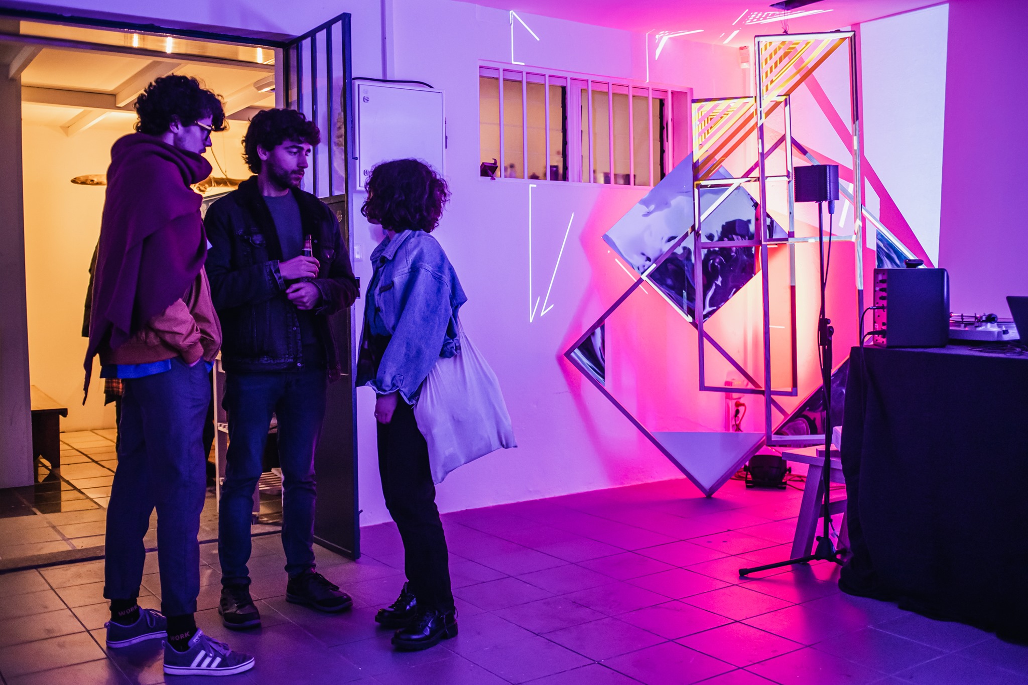 Troje ludzi, dwóch mężczyzn i kobieta stoją na korytarzu i rozmawiają, obok nich stoi podświetlona geometryczna konstrukcja, we fragment której wpasowano abstrakcyjny obraz.