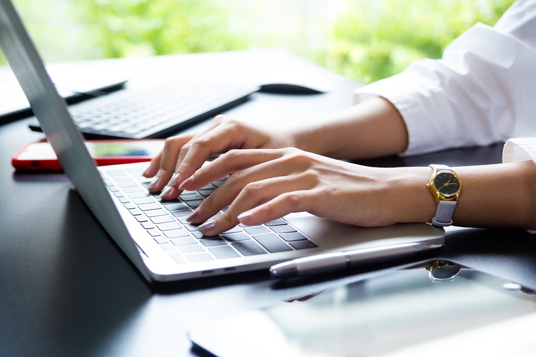 zbliżenie na kobiece dłonie piszące po klawiaturze laptopa. Na nadgarstku widać zegarek, a paznokcie się pomalowane na fioletowo