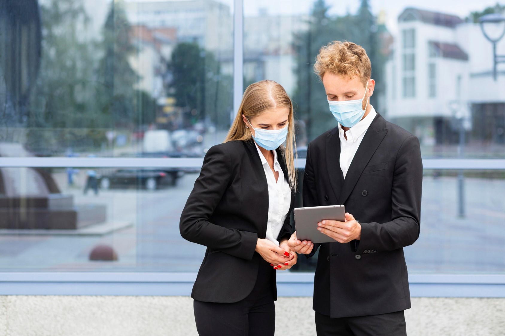 Młodzi ludzie: kobieta i mężczyzna stoją na tle szklanego budynku, mężczyzna pokazuje coś kobiecie na tablecie. Obydwoje mają maseczki chirurgiczne zakrywające usta i nos.